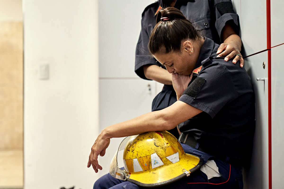 Female firefighter in distress sits with head bowed, comforted by a colleague, helmet in lap. Signifies need for trauma therapy.